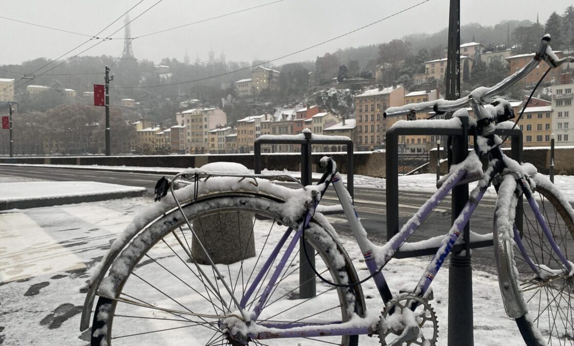 Un vélo enneigé au premier plan, sur les quais de saone à Lyon. Au loin, on distingue fourvière.